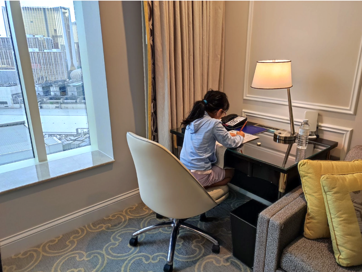 旅行中のホテル客室で朝の宿題に取り組む子ども。窓外の街並みを背景に、学習習慣を続ける旅育の様子