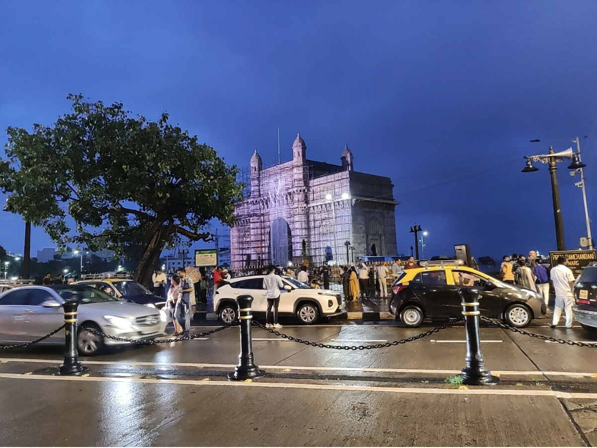 ムンバイのランドマーク「インド門」。夕暮れの雨上がり、観光客と車で賑わう海沿いの広場の風景