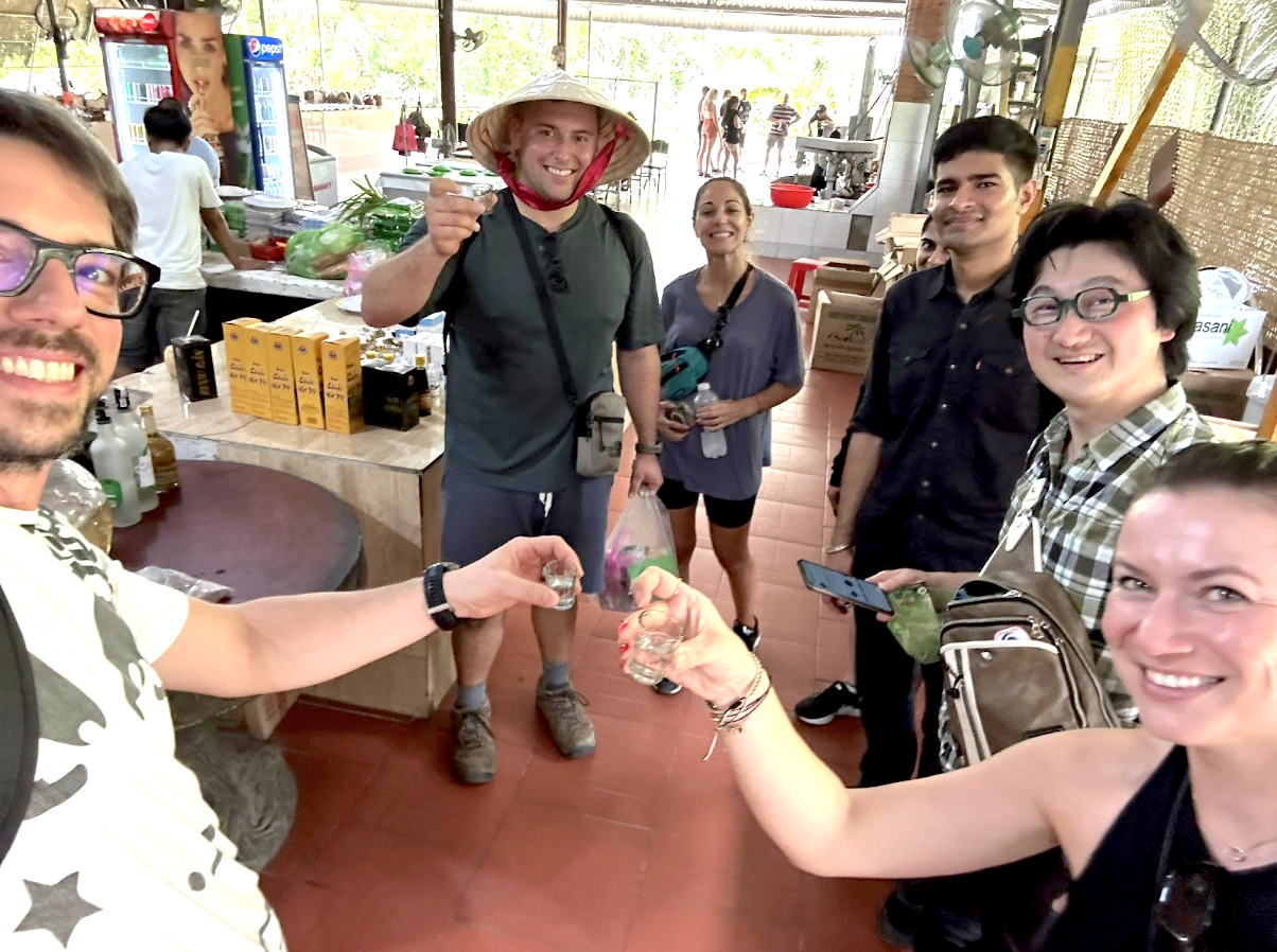 ベトナム・ホーチミンのローカル市場で、ツアー仲間と小さなグラスで乾杯する旅行者たち。交流を楽しむ明るい雰囲気の一枚